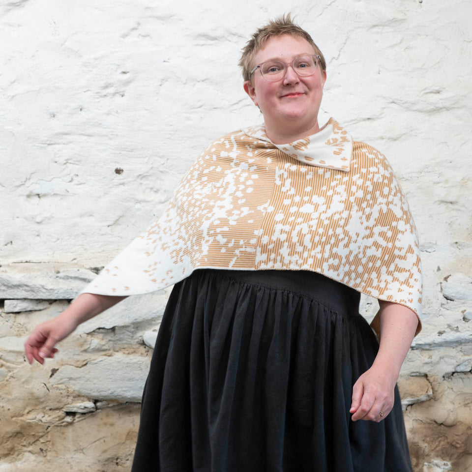 Felix Ford stands in a rustic, whitewashed stone shed in Hoswick, Shetland. A white woman with short, fair hair wears spectacles and - over a black linen dress with gathered skirt - a finely knitted cape in a speckled pattern in cream, gold and brown. She looks at the camera, smiles, and dances with her arms out to her sides.