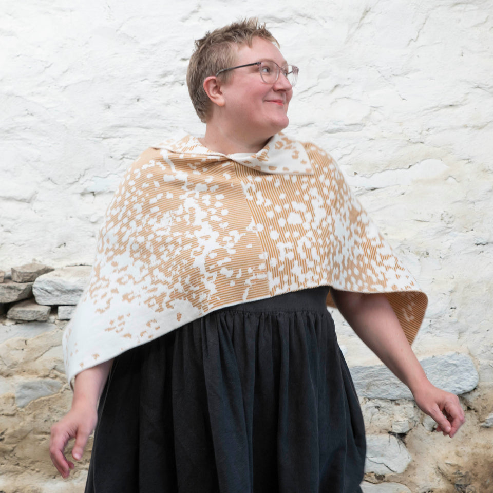Felix Ford stands in a rustic, whitewashed stone shed in Hoswick, Shetland. A white woman with short, fair hair wears spectacles and - over a black linen dress with gathered skirt - a finely knitted cape in a speckled pattern in cream, gold and brown. She away from the camera, smiles, and dances with her arms out to her sides.