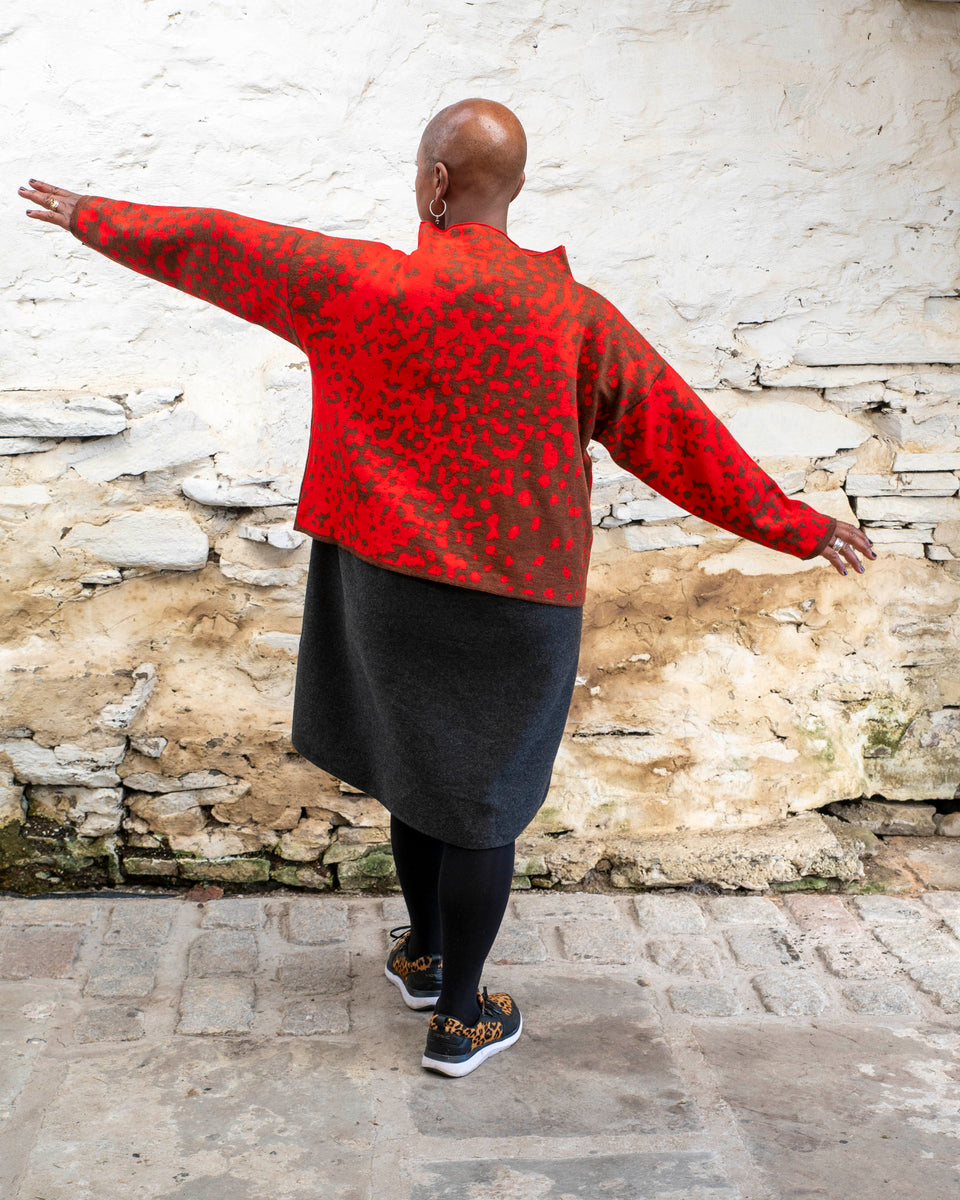 Jeanette Sloan, a black woman with a shaved head stands inside a rustic, whitewashed, stone building in Hoswick Shetland. She is wearing largish, silver hooped earrings with a bead hanging, a tomato red and chestnut brown finely knitted boxy jumper with a graphic animal print patter and a high neck, a carcoal grey woolen dress, black opaque tights and black leather and leopard print trainers. She is standing with her back to the camera and holds her arms out to show the shape of the jumper.
