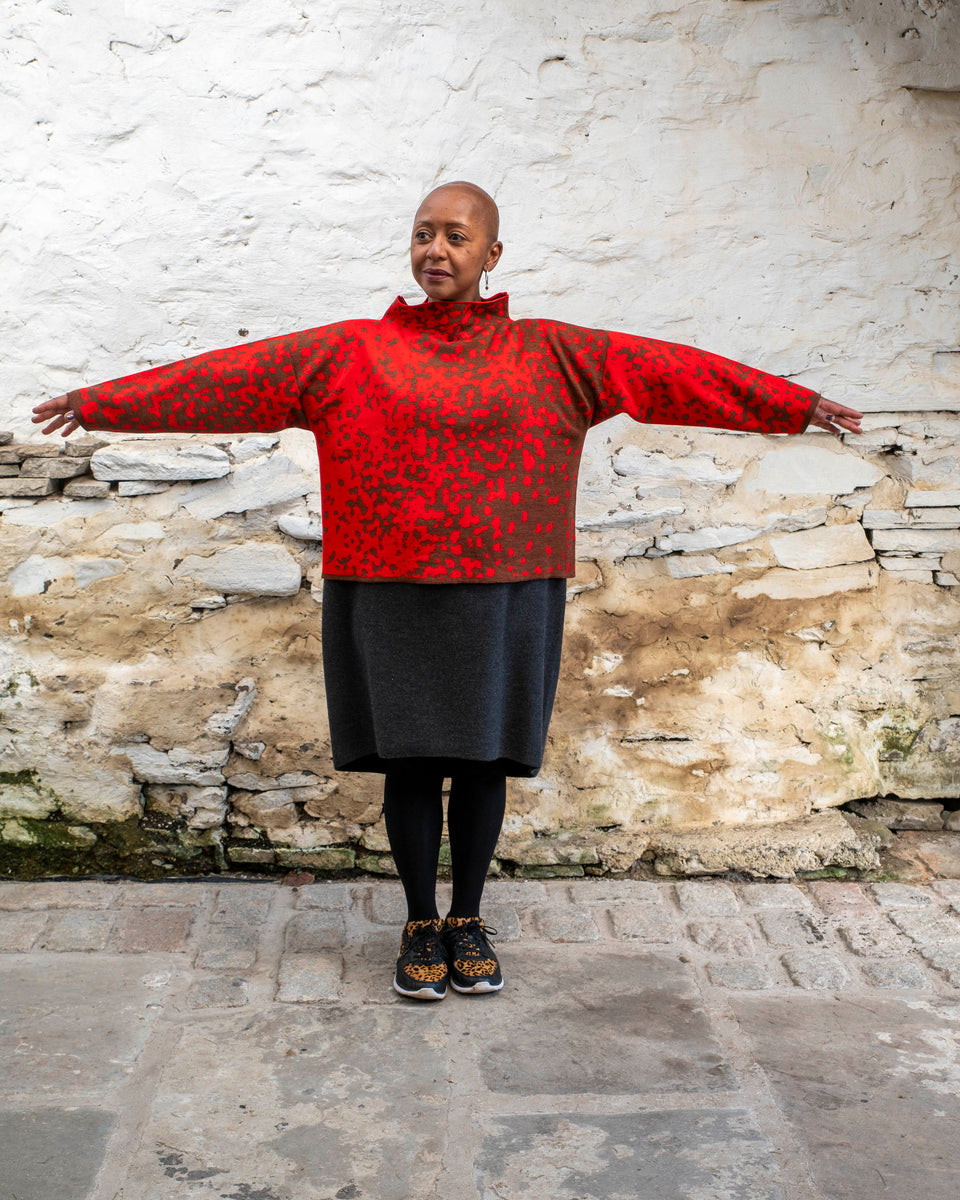 Jeanette Sloan, a black woman with a shaved head stands inside a rustic, whitewashed, stone building in Hoswick Shetland. She is wearing largish, silver hooped earrings with a bead hanging, a tomato red and chestnut brown finely knitted boxy jumper with a graphic animal print patter and a high neck, a carcoal grey woolen dress, black opaque tights and black leather and leopard print trainers. She is standing straight on with her arms held our to show the shape of the jumper. Shel looks to her right.