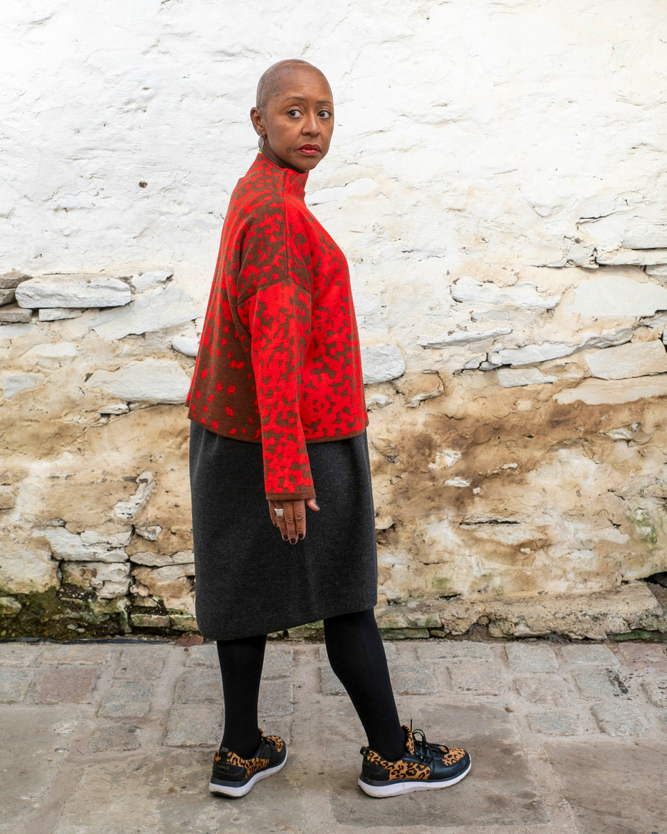 Jeanette Sloan, a black woman with a shaved head stands inside a rustic, whitewashed, stone building in Hoswick Shetland. She is wearing largish, silver hooped earrings with a bead hanging, a tomato red and chestnut brown finely knitted boxy jumper with a graphic animal print patter and a high neck, a carcoal grey woolen dress, black opaque tights and black leather and leopard print trainers. She is standing with her right side to the camera and looks to her left.