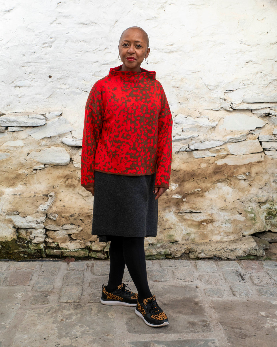 Jeanette Sloan, a black woman with a shaved head stands inside a rustic, whitewashed, stone building in Hoswick Shetland. She is wearing largish, silver hooped earrings with a bead hanging, a tomato red and chestnut brown finely knitted boxy jumper with a graphic animal print patter and a high neck, a carcoal grey woolen dress, black opaque tights and black leather and leopard print trainers. She looks forward and smiles at the camera.