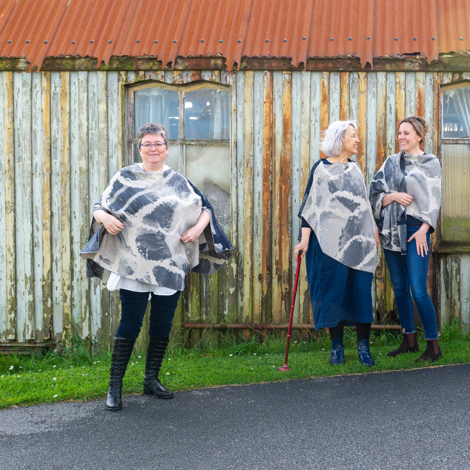 A white woman with short brown and grey hair stands in front of an old timber building with peeling paint in Hoswick, Shetland. She is wearing purple, metal rimmed glasses, navy velvet leggings tucked into black leather knee high boots and a finely knitted navy and off white cape in an abstract pattern. Underneath is a mens white, dress shirt. Next to her two white women, one with grey hair and the other with brown hair. Both wearing denim and finely knitted knitwear in navy and off white merino.