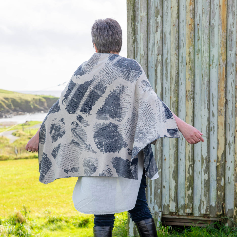 A white woman with short brown and grey hair stands in front of an old timber building with peeling paint in Hoswick, Shetland. She is wearing purple, metal rimmed glasses, navy velvet leggings tucked into black leather knee high boots and a finely knitted navy and off white cape in an abstract pattern. Underneath is a mens white, dress shirt. She stands with her back to the camera and the cape flies out. It's a sunny day, with hoswick bay in the background.
