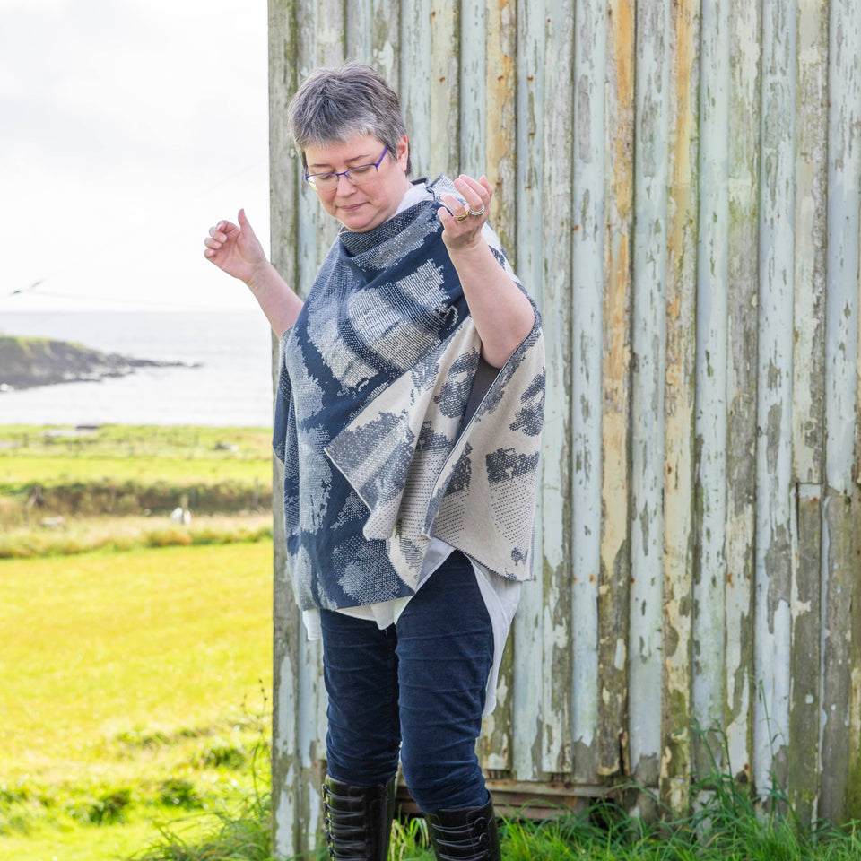 A white woman with short brown and grey hair stands in front of an old timber building with peeling paint in Hoswick, Shetland. She is wearing purple, metal rimmed glasses, navy velvet leggings tucked into black leather knee high boots and a finely knitted navy and off white cape in an abstract pattern. Underneath is a mens white, dress shirt.