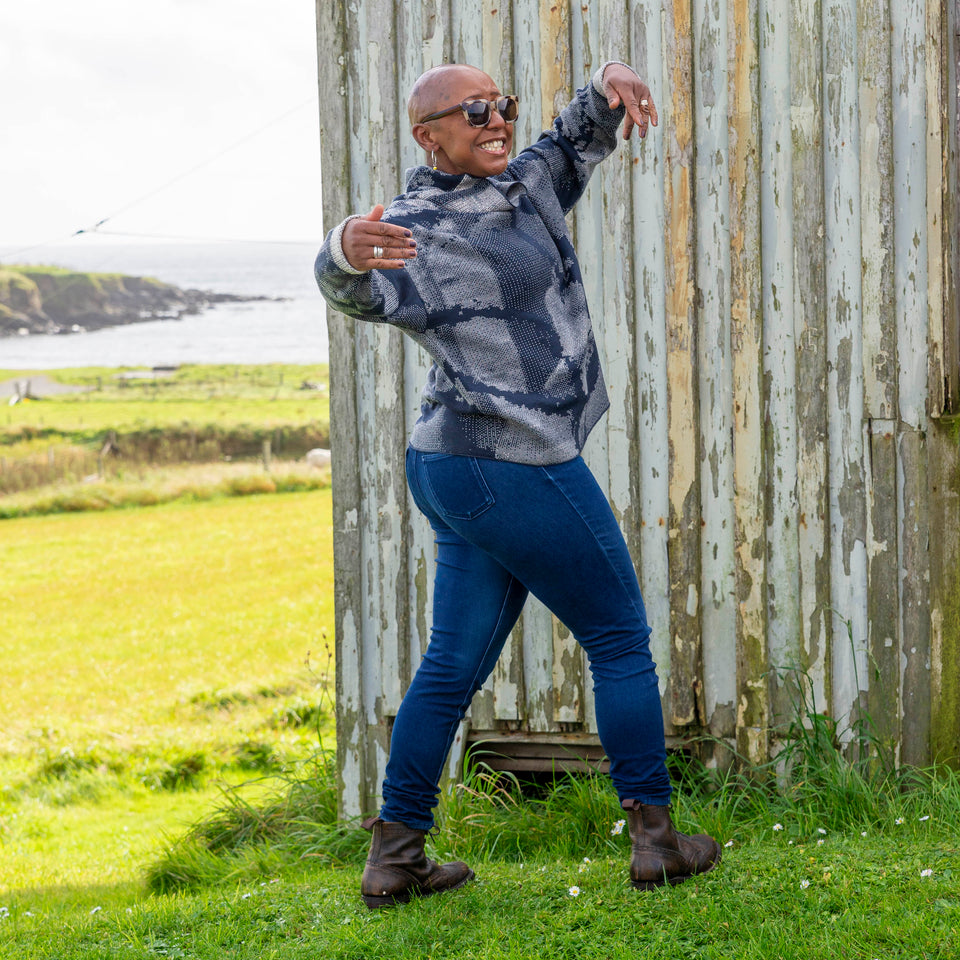 Jeanette Sloan at Hoswick, Shetland. She stands on the grass with sea behind. She wears tortoiseshell sunglasses, hoop earrings, a cowl necked Nielanell jumper in navy and off white abstract pattern, dark indigo jeans and brown lace up boots.