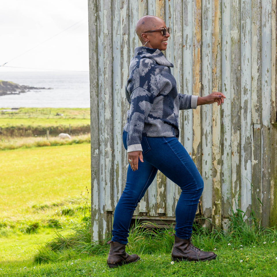 Jeanette Sloan at Hoswick, Shetland. She stands on the grass with sea behind. She wears tortoiseshell sunglasses, hoop earrings, a cowl necked Nielanell jumper in navy and off white abstract pattern, dark indigo jeans and brown lace up boots.