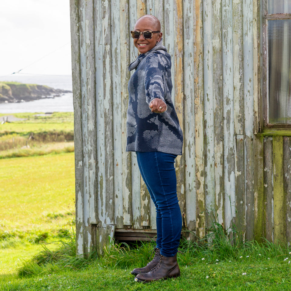 Jeanette Sloan at Hoswick, Shetland. She stands on the grass with sea behind. She wears tortoiseshell sunglasses, hoop earrings, a cowl necked Nielanell jumper in navy and off white abstract pattern, dark indigo jeans and brown lace up boots.