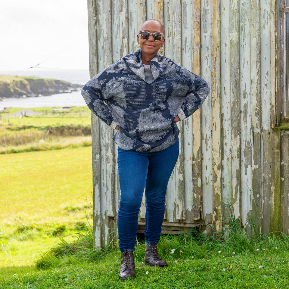 Jeanette Sloan at Hoswick, Shetland. She stands on the grass with sea behind. She wears tortoiseshell sunglasses, hoop earrings, a cowl necked Nielanell jumper in navy and off white abstract pattern, dark indigo jeans and brown lace up boots.
