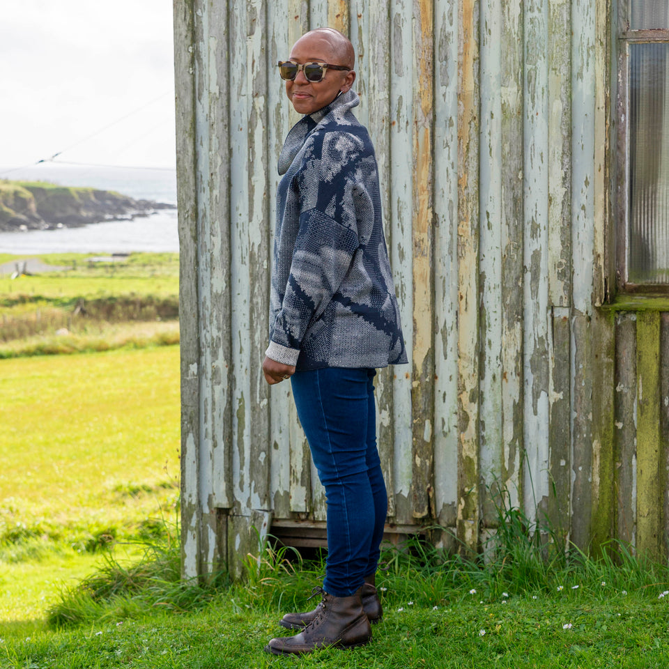 Jeanette Sloan at Hoswick, Shetland. She stands on the grass with sea behind. She wears tortoiseshell sunglasses, hoop earrings, a cowl necked Nielanell jumper in navy and off white abstract pattern, dark indigo jeans and brown lace up boots.