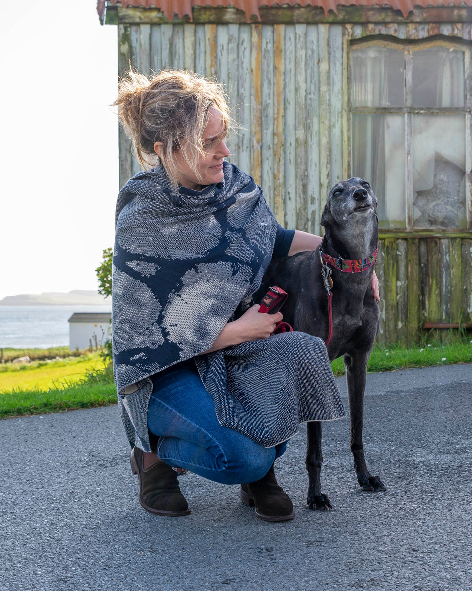 A white woman with fair hair tied back crouches beside a black greyhound outside the old Gospel Hall. Hoswick. She is wearing a contemporary shawl, in navy and off white, wrapped around her shoulders. She also wears a blue long sleeve t shirt, blue jeans and brown suede dealer boots.