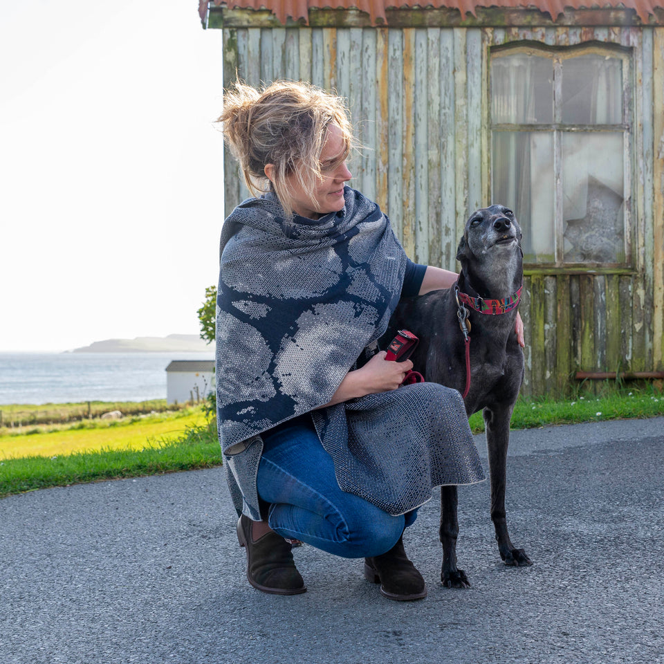A white woman with fair hair tied back crouches beside a black greyhound outside the old Gospel Hall. Hoswick. She is wearing a contemporary shawl, in navy and off white, wrapped around her shoulders. She also wears a blue long sleeve t shirt, blue jeans and brown suede dealer boots.