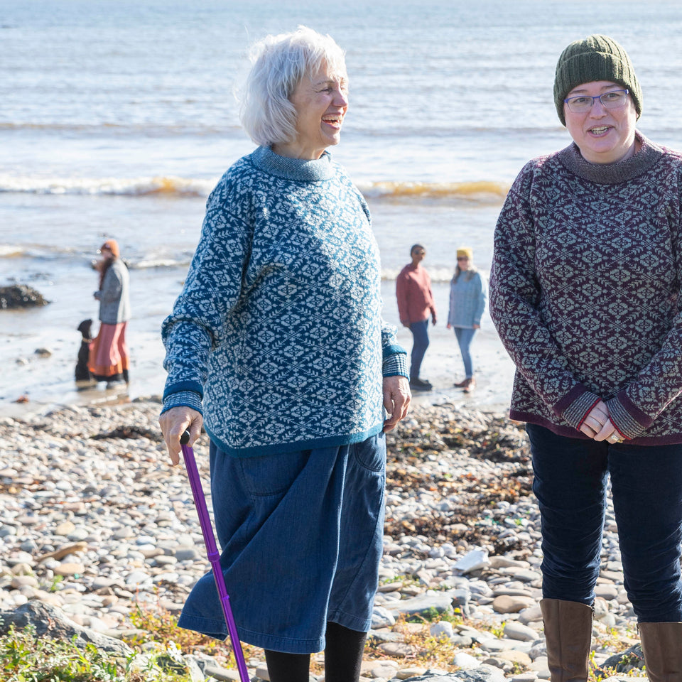 A woman with jaw length grey hair wears a blue and grey fair isle patterned jumper over a loose denim dress. She is side on to the camera and has a purple walking stick in her right hand. To her right another woman in a wine and leichen coloured fair isle jumper. She is wearing jeans and knee lenght brown leather boots and a dark green ribbed beanie hat.. In the background is the sea at Hoswick. On the shore a young woman with a black labrador and two women milling about wearing jumpers.