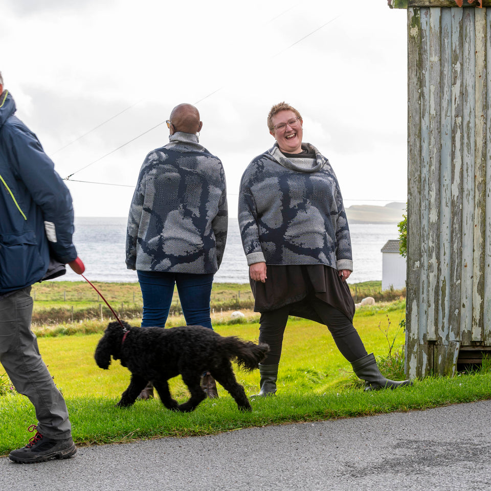 One black and one white woman on the grass at Hoswick, Shetland with the sea behind. They are both wearing the same cowl-necked contemporary merino jumper. A contemporary Shetland sweater in navy and off white with an abstract pattern.