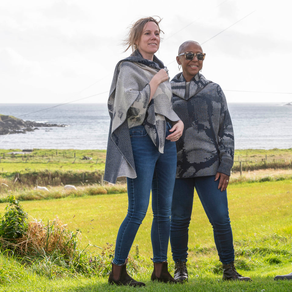 A white woman with fair hair tied back and a black woman with a shaved head stand on the grass in fields above Hoswick bay, Shetland. They are both wearing contemporary knitwear in navy and off white. On the left the woman wears a contempoary shawl wrapped around her and on the right the woman wears a cowl necked jumper. Both wear jeans and brown boots.