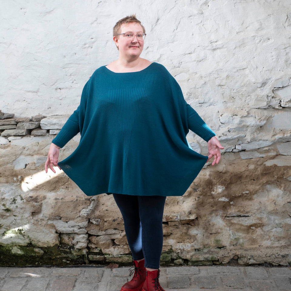 A white woman with short fair hair stands in a rustic stone shed. She faces the camera and looks straight ahead. She is wearing a loose and finely knit jumper in dark teal over navy leggings and red chunky lace up boots. She holds the bottom edges of the sweater out to show its shape.