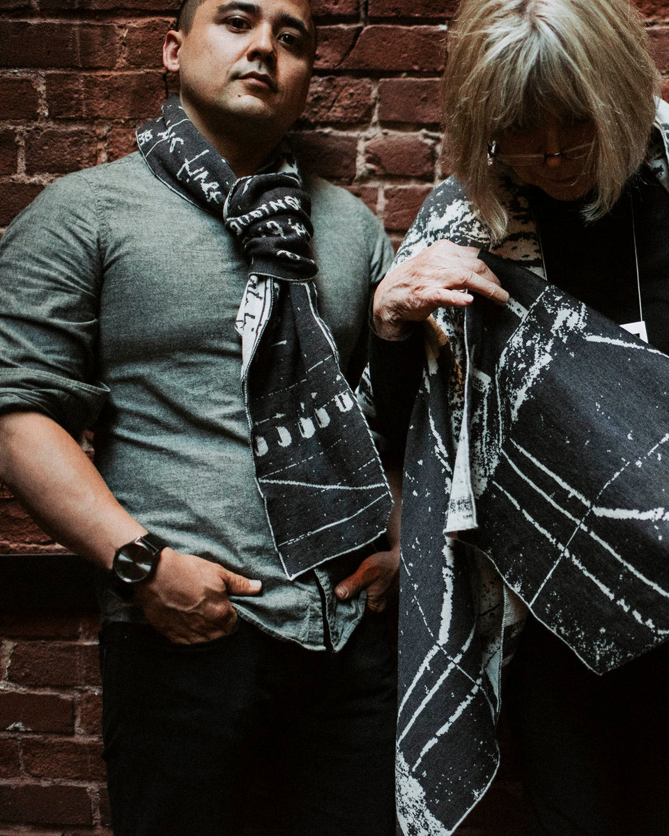 A man and woman wear contemporary art-knitwear in charcoal and stone white. They stand against a brick wall.