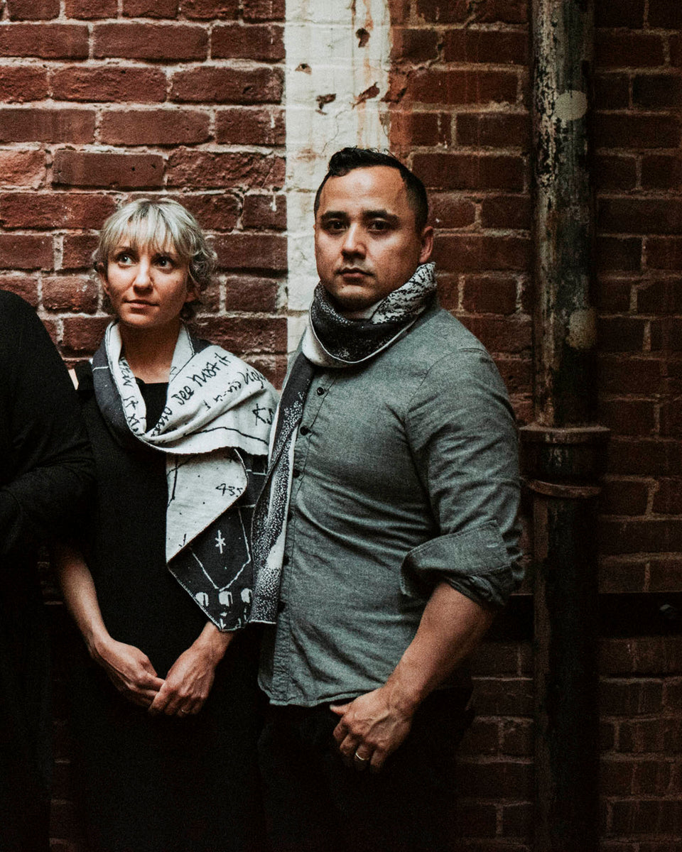 A man and woman wear contemporary art-knitwear in charcoal and stone white. They stand against a brick wall.