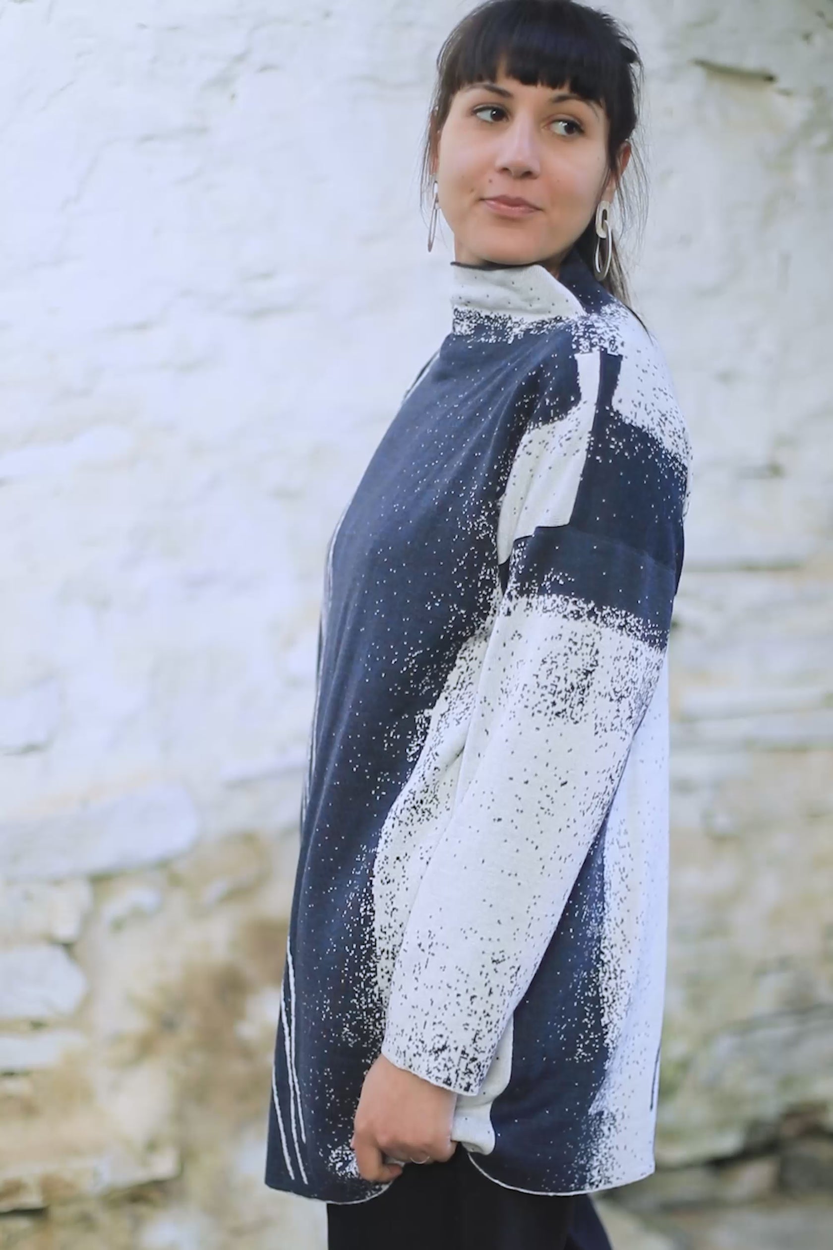 Video shot inside a small, rustic stone building in Hoswick, Shetland. A tall woman with olive skin and dark, straight brown hair (worn tied back in a high ponytail) wears a longline midnight blue and pale grey jumper with an abstracted motif evocative of the sea shore. Lines much like rivulets made by water, sand speckles.