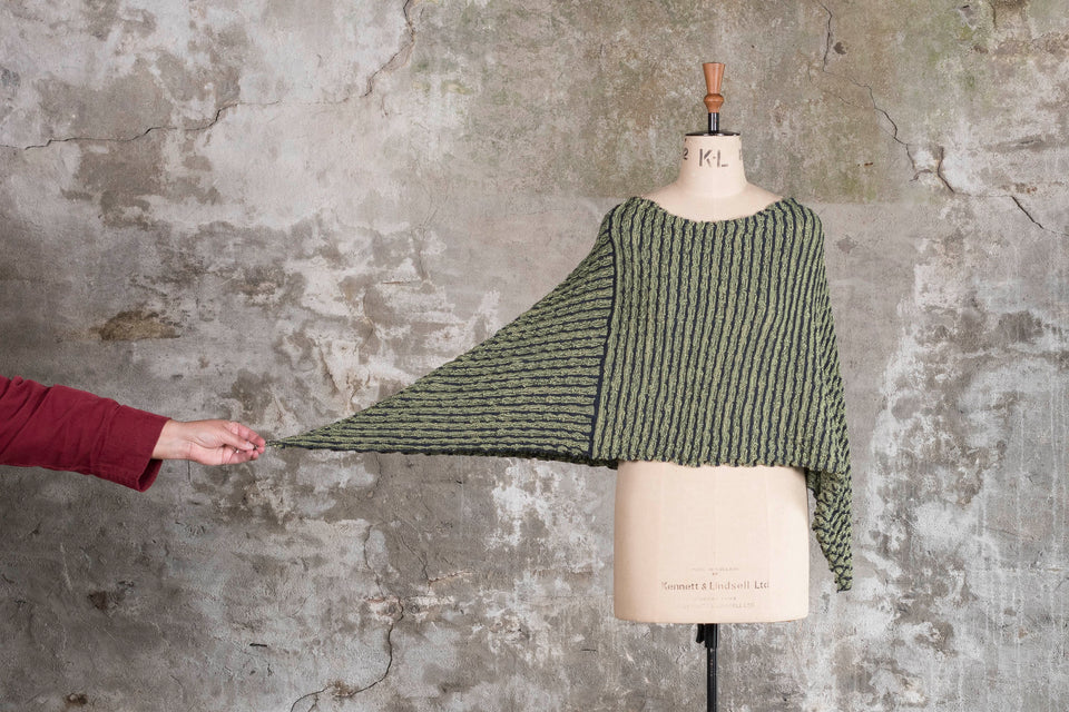 Knitted rigg cape on mannequin, with side being pulled out to show shape. The textile is ridged and with stripes of colour knitted in. Shown here in a dark soft green and charcoal