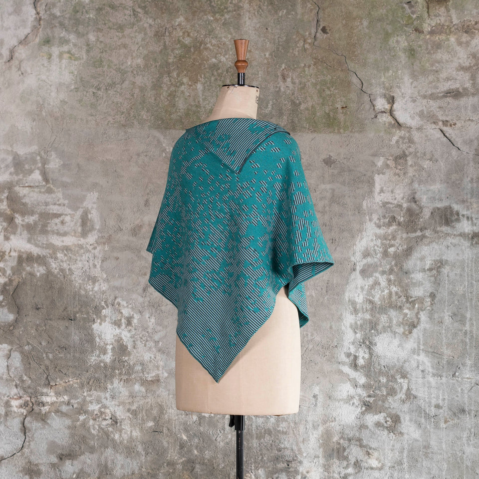 On a vintage mannequin against a rustic, old plastered wall, is a finely knitted cape with a mottled and striped pattern. There are two teals, one light and the other dark, with a very dark blue fine stripe. Shown from the back and turned slightly towards the right shoulder.