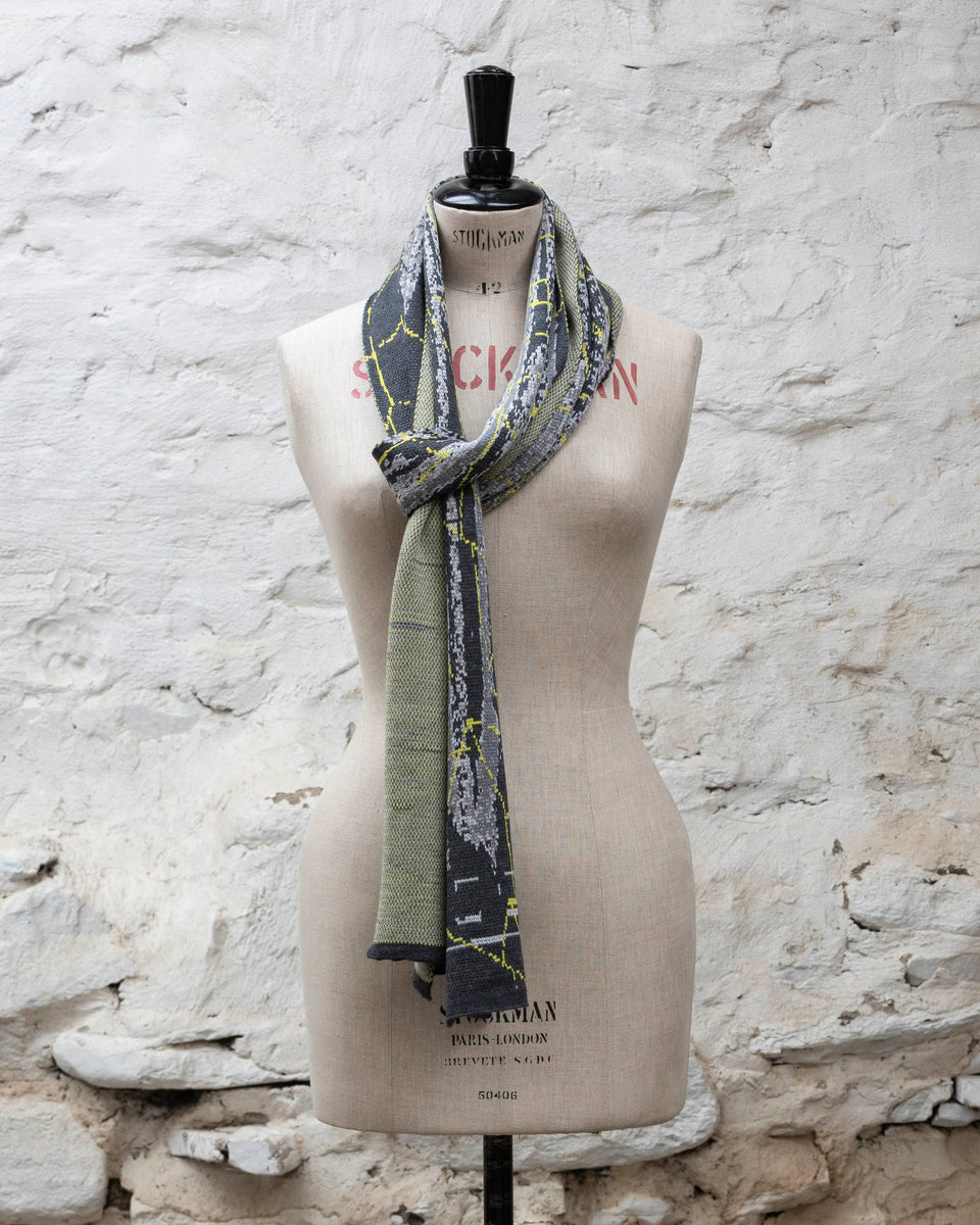 A grey scarf with abstract patterning and linear limey yellow  highlights. Shown in a loose Parisian scarf knot. Large and luxurious scarf knitted in fine merino. Shown on a vintage mannequin against a rustic whitewashed wall