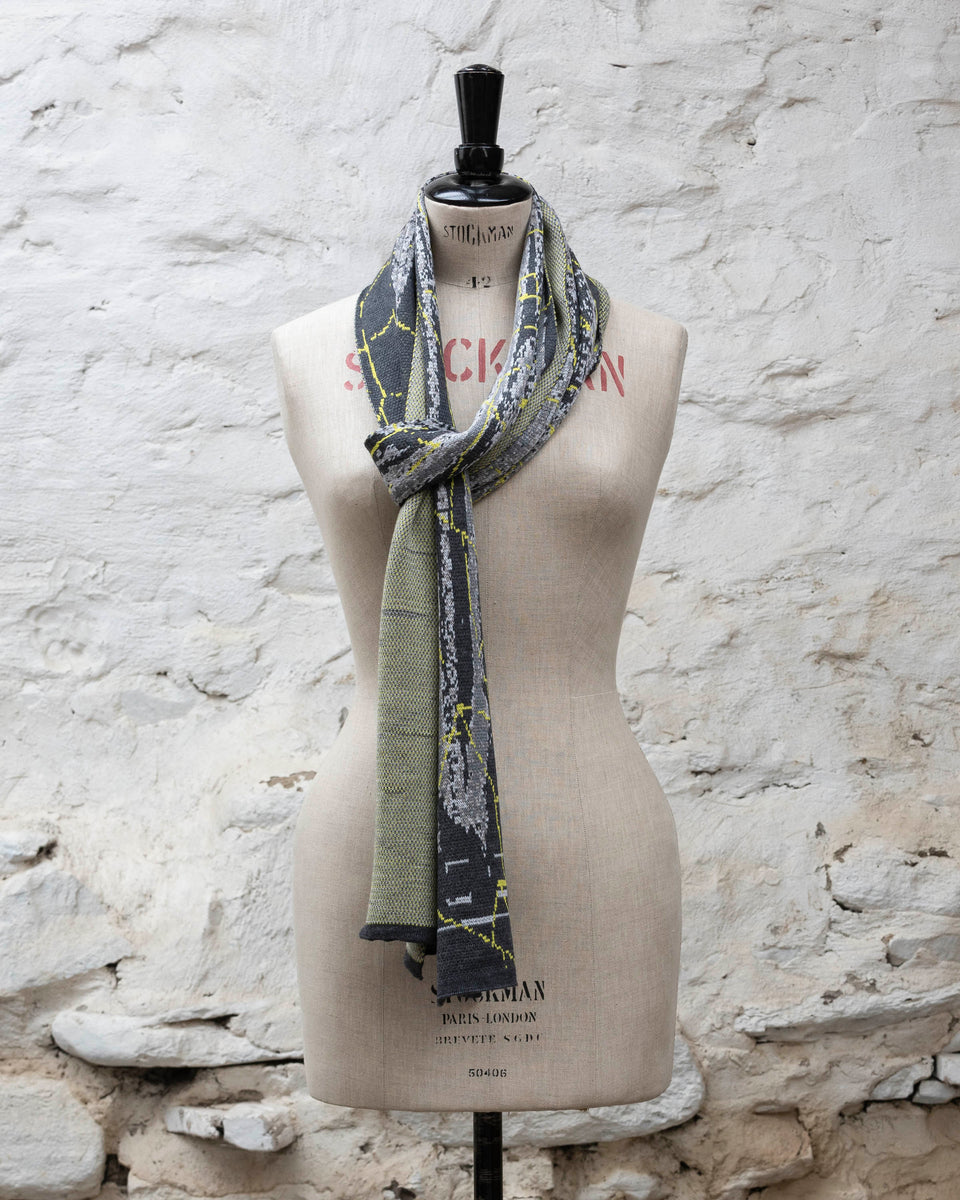 A grey scarf with abstract patterning and linear limey yellow highlights. Shown in a loose Parisian scarf knot. Large and luxurious scarf knitted in fine merino. Shown on a vintage mannequin against a rustic whitewashed wall