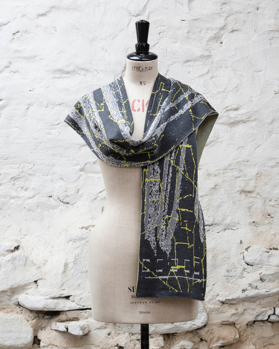 A grey scarf with abstract patterning and linear limey yellow highlights. Shown loosely draped with one end over shoulder. Large and luxurious scarf knitted in fine merino. Shown on a vintage mannequin against a rustic whitewashed wall