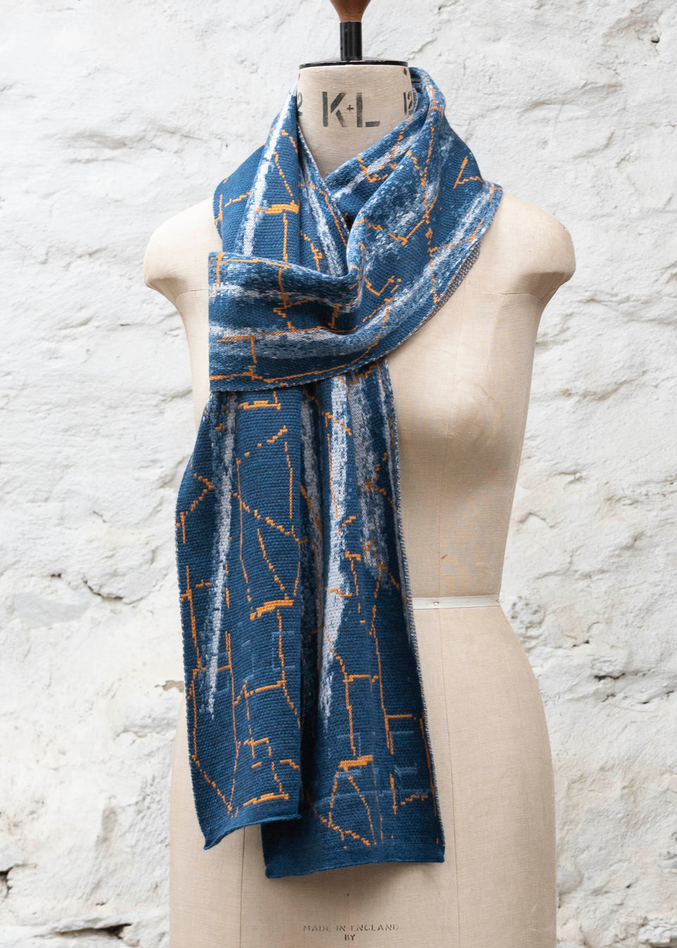A blue scarf with abstract patterning and linear orange highlights. Shown in the Parisian scarf knot. Large and luxurious scarf knitted in fine merino. Shown on a vintage mannequin against a rustic whitewashed wall
