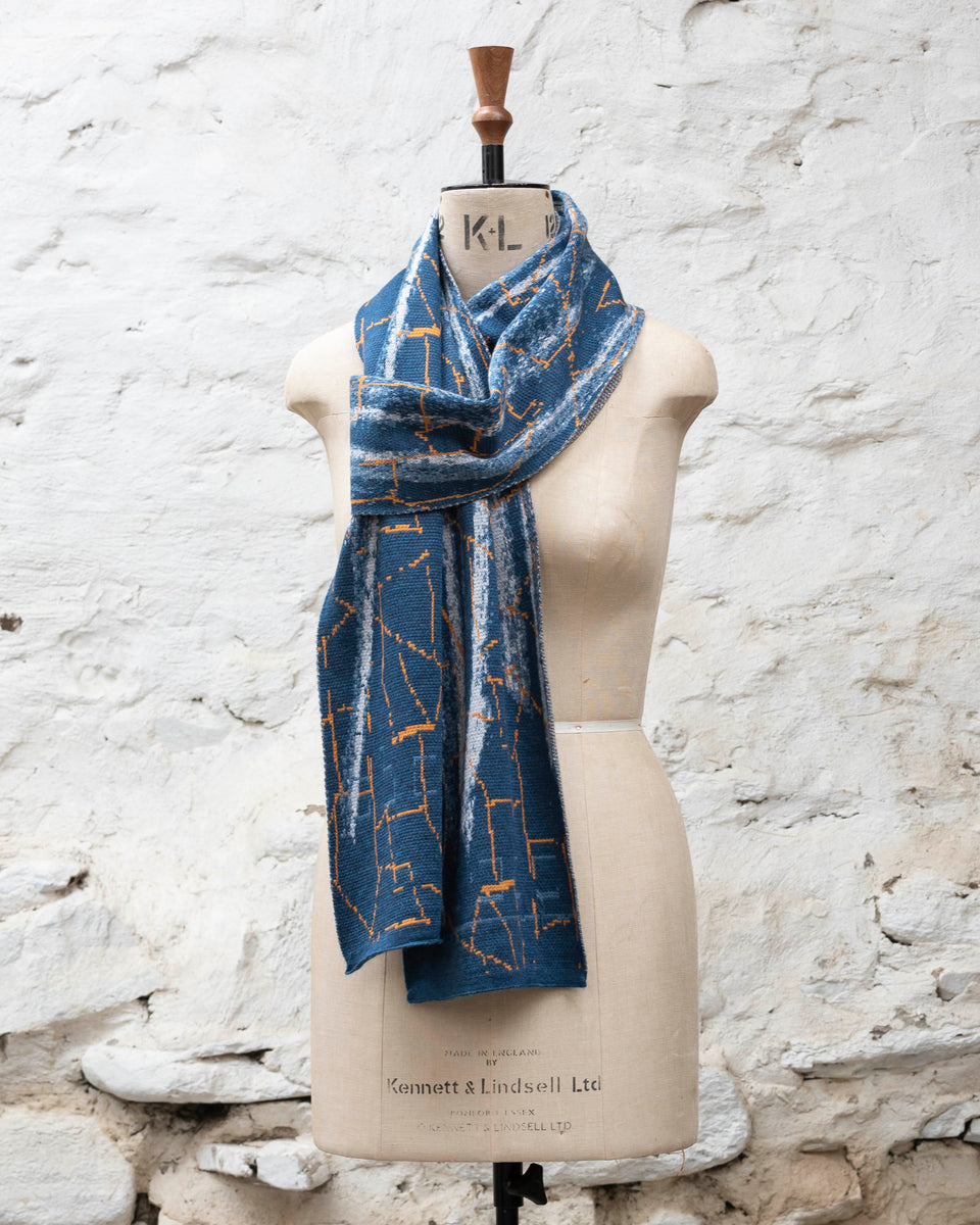A blue scarf with abstract patterning and linear orange highlights. Shown in the Parisian scarf knot. Large and luxurious scarf knitted in fine merino. Shown on a vintage mannequin against a rustic whitewashed wall