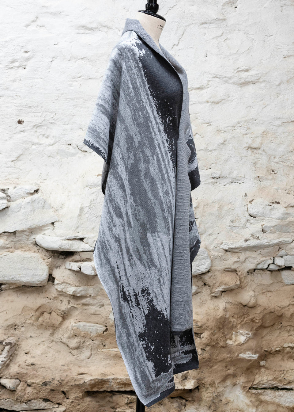 Finely knitted shawl in luxurious merino, made in Scotland. Shown on a vintage mannequin against a whitewashed wall. Abstract design in sea greys with curvilinear motifs. Draped to show whole shawl from theside