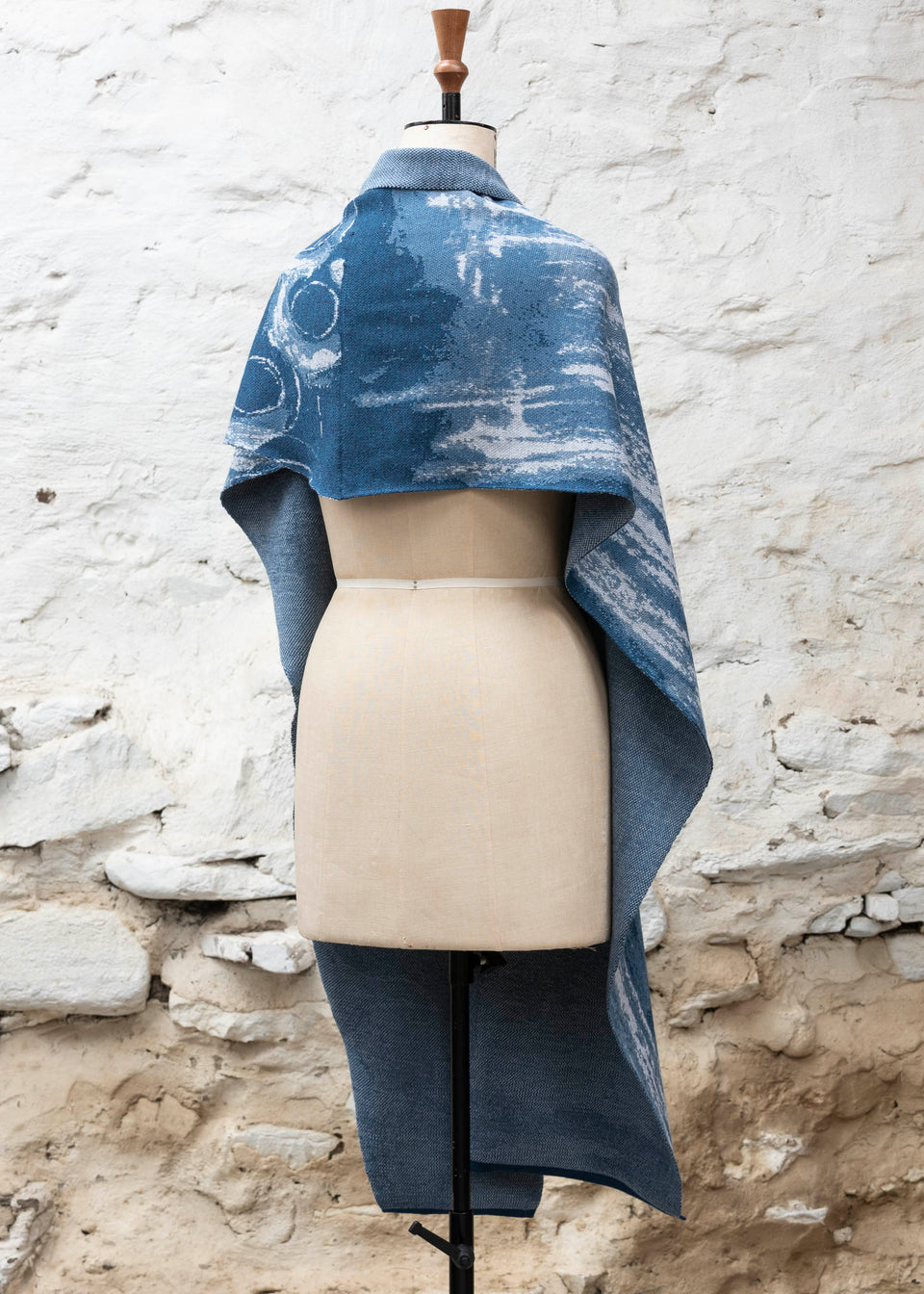 Finely knitted shawl in luxurious merino, made in Scotland. Shown on a vintage mannequin against a whitewashed wall. Abstract design in sea blues with curvilinear motifs. Draped to show full wrap from the back