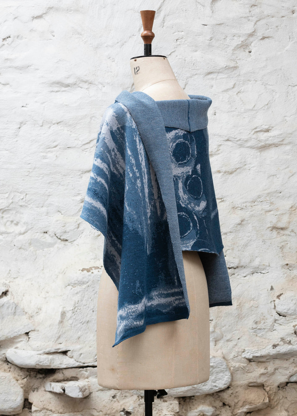 Finely knitted shawl in luxurious merino, made in Scotland. Shown on a vintage mannequin against a whitewashed wall. Abstract design in sea blues with curvilinear motifs. Wrapped around for a shorter style and shown from side