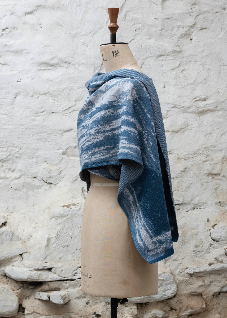 Finely knitted shawl in luxurious merino, made in Scotland. Shown on a vintage mannequin against a whitewashed wall. Abstract design in sea blues with curvilinear motifs. Wrapped around for a shorter style and shown from side