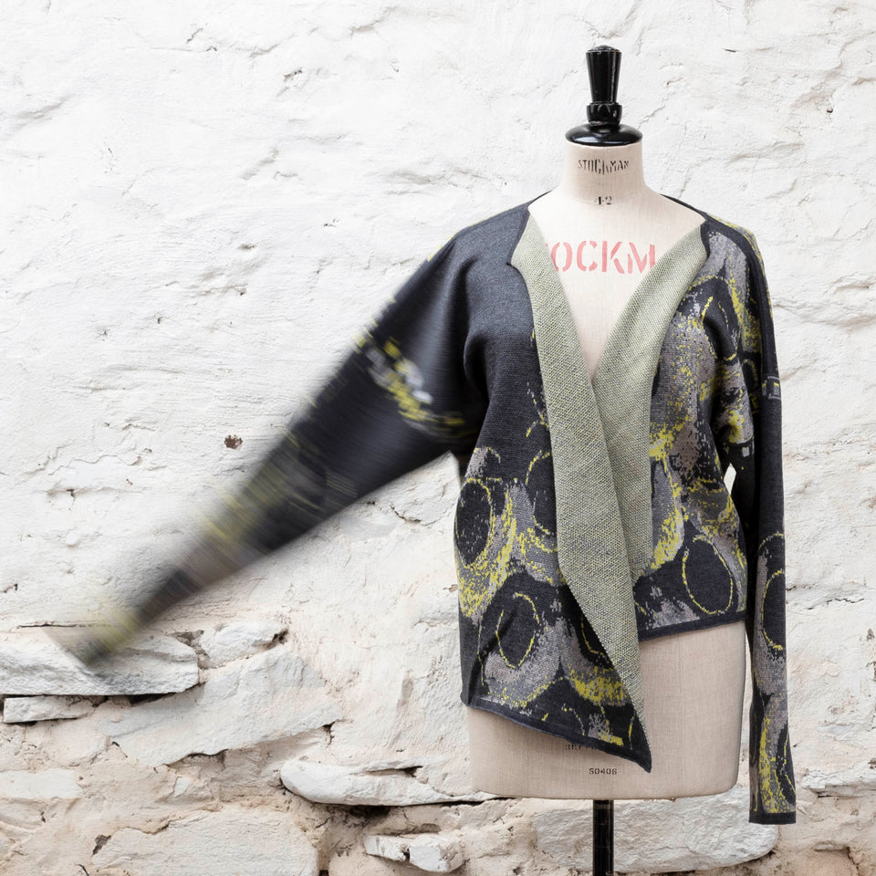 Knitted jacket inspired by the Shetland coastal village of Hoswick. Abstract curvilinear motif in greys and fluoro yellow. Asymmetric design with lapels that fold back. Shown front-on, on a vintage mannequin, against a white washed rustic stone wall