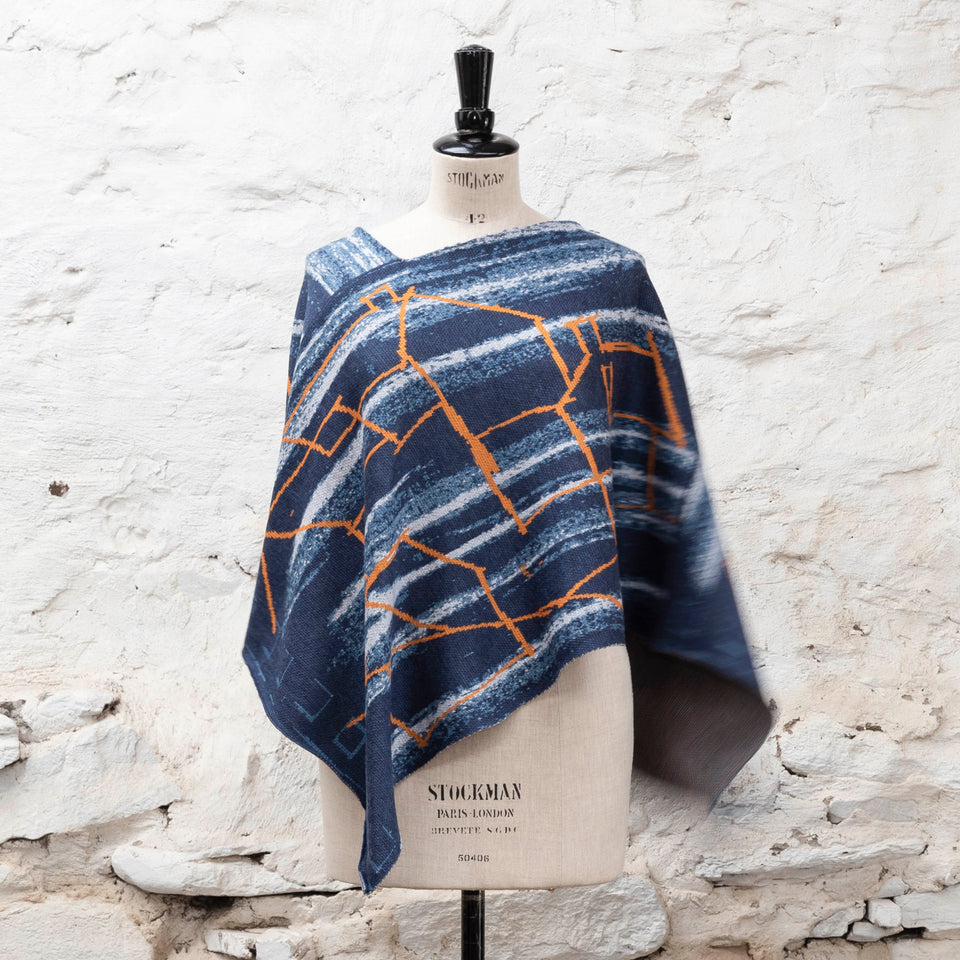 A knitted poncho, made in Shetland in a contemporary design by Nielanell with abstract blues and orange linear marks. Shown front on, on a vintage mannequin against a rustic white washed wall