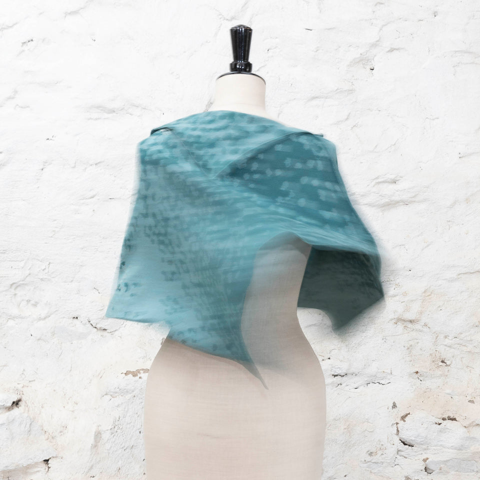On a vintage mannequin and shown against a rustic, whitewashed stone wall a teal cape swings. It is blurred with the movement and the pattern is two tone with a mottled and striped effect.