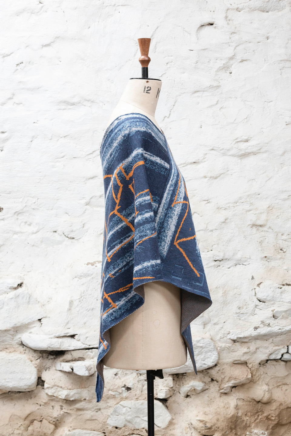 A knitted poncho, made in Shetland in a contemporary design by Nielanell with abstract blues and orange linear marks. Shown exactly side on, on a vintage mannequin against a rustic white washed wall