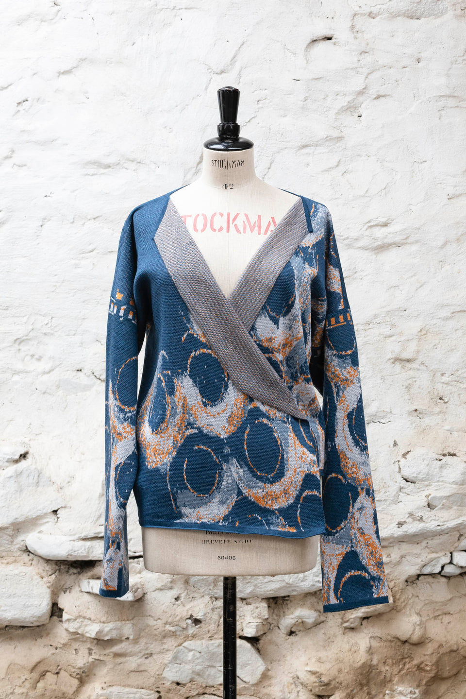 Knitted jacket inspired by the Shetland coastal village of Hoswick. Abstract curvilinear motif in blues with orange highlights . Asymmetric design with lapels that fold back. Shown front-on, on a vintage mannequin, against a white washed rustic stone wall