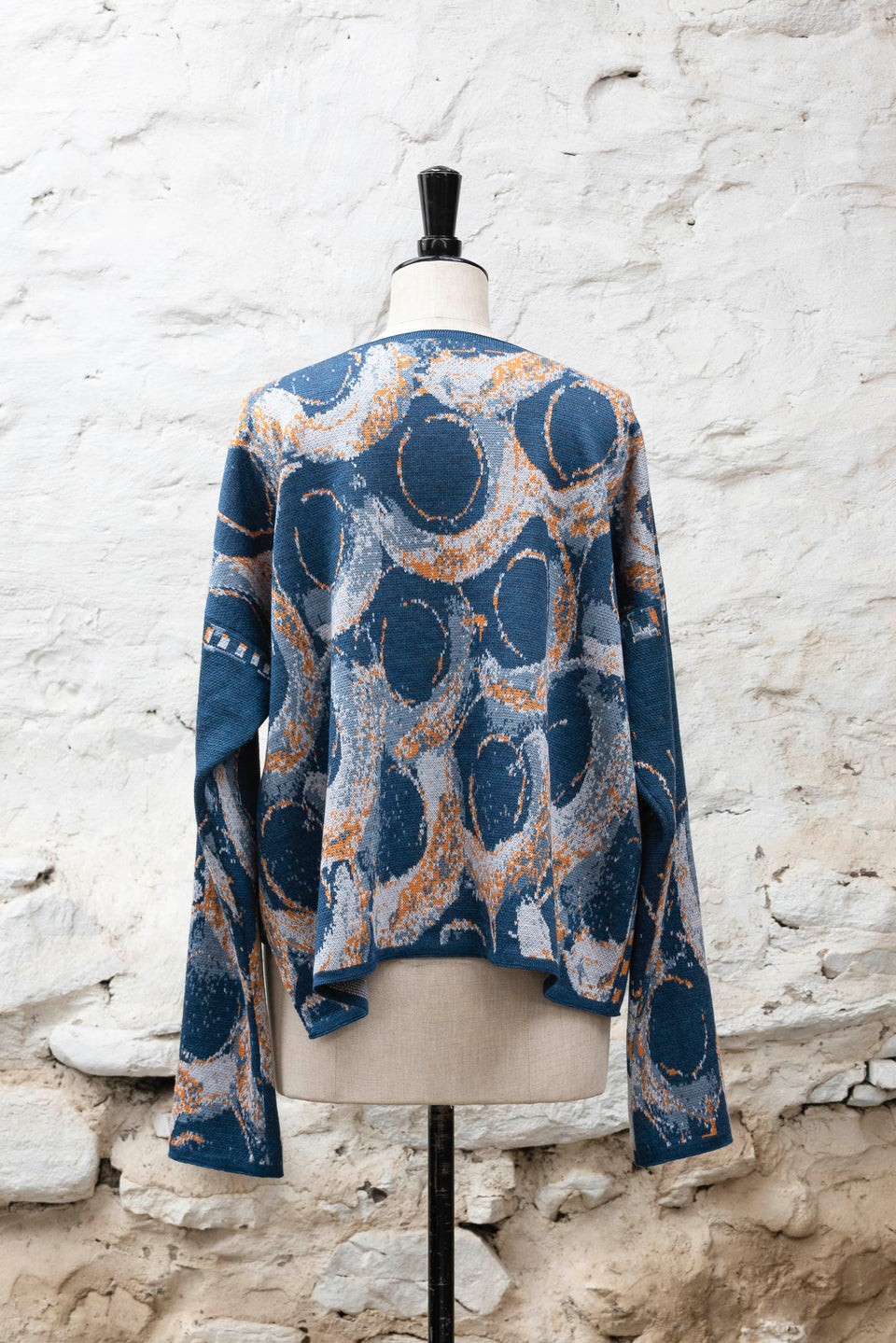 Knitted jacket inspired by the Shetland coastal village of Hoswick. Abstract curvilinear motif in blues with orange highlights . Asymmetric design with lapels that fold back. Shown from the back, on a vintage mannequin, against a white washed rustic stone wall