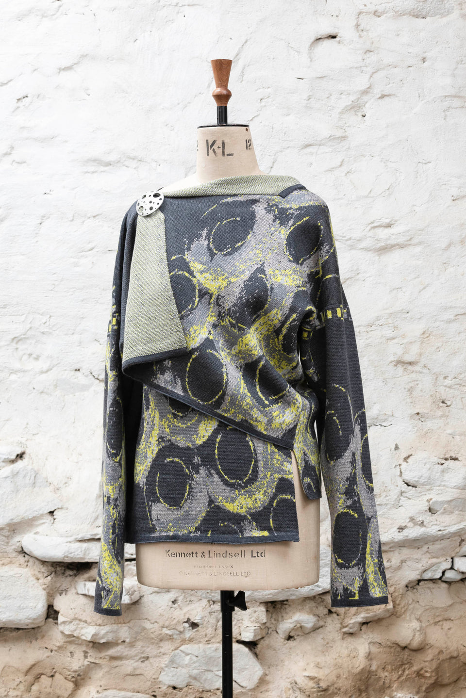 Knitted jacket inspired by the Shetland coastal village of Hoswick. Abstract curvilinear motif in greys and fluoro yellow. Asymmetric design with lapels that fold back. Shown front-on, on a vintage mannequin with body pinned at shoulder with a round brooch., against a white washed rustic stone wall