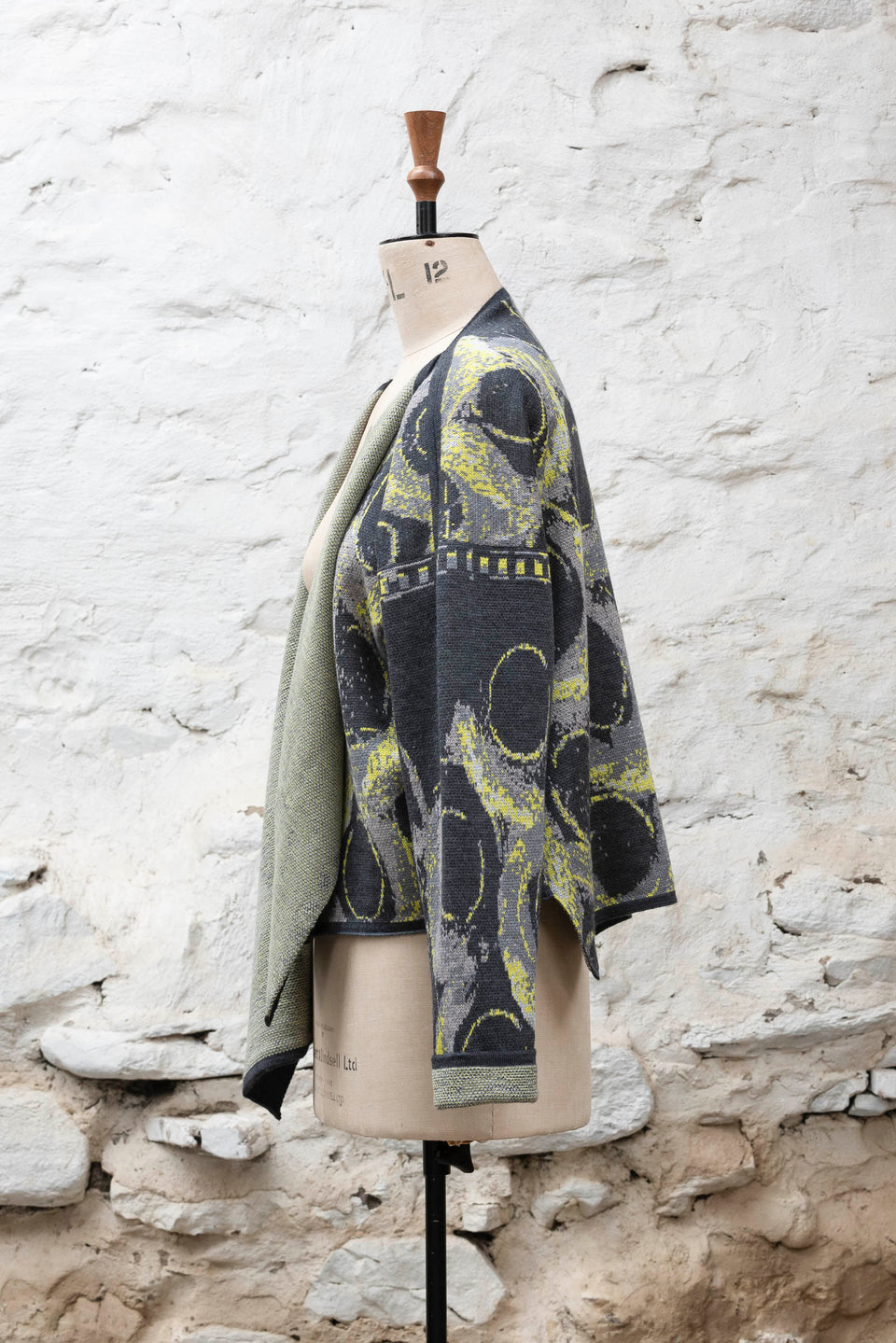 Knitted jacket inspired by the Shetland coastal village of Hoswick. Abstract curvilinear motif in greys and fluoro yellow. Asymmetric design with lapels that fold back. Shown side-on, on a vintage mannequin, against a white washed rustic stone wall
