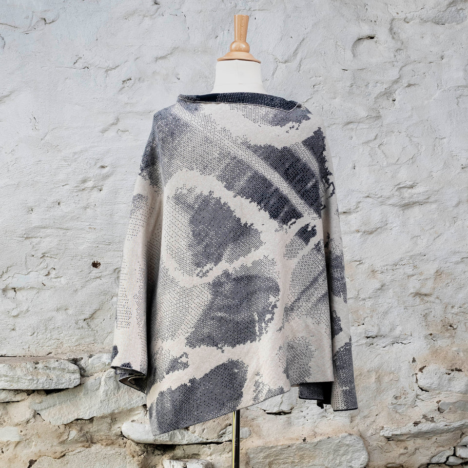 Knitted Rani cape. Small abstract patterns make up a larger design with photographic imagery. Shown in inky blue and antique white