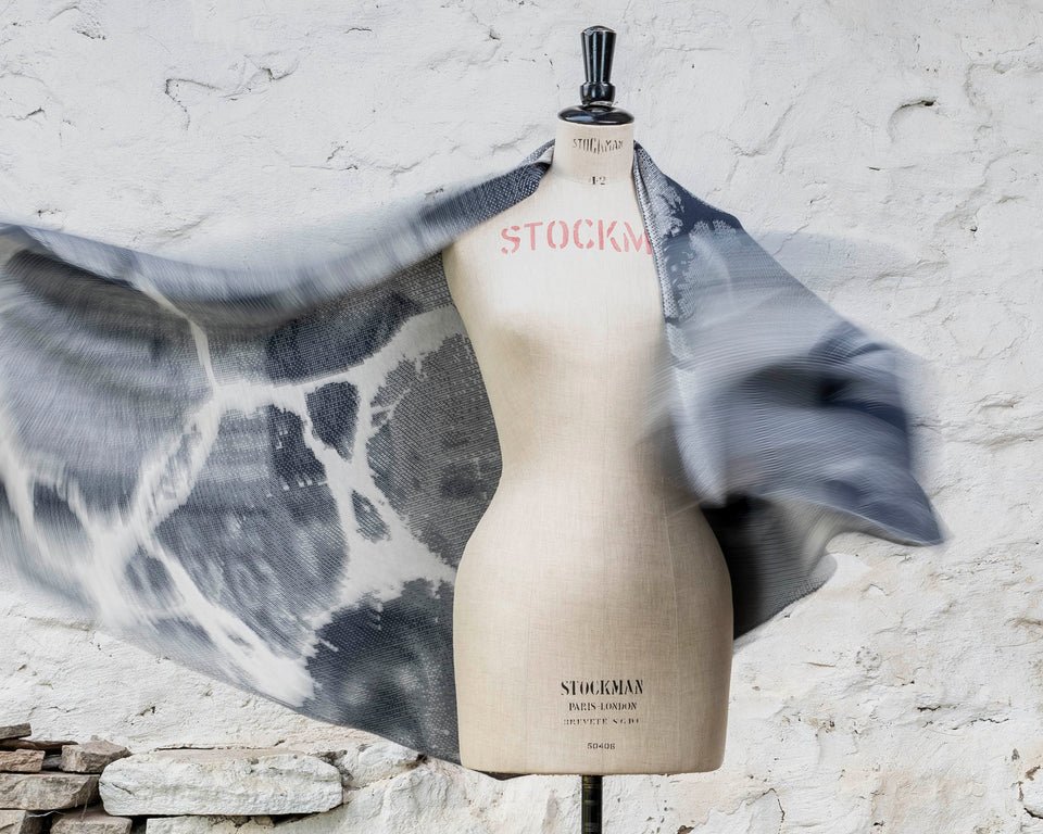 Knitted Rani shawl / wrap. Small abstract patterns make up a larger design with photographic imagery. Shown on mannequin with shawl flying out. Inky blue and antique white