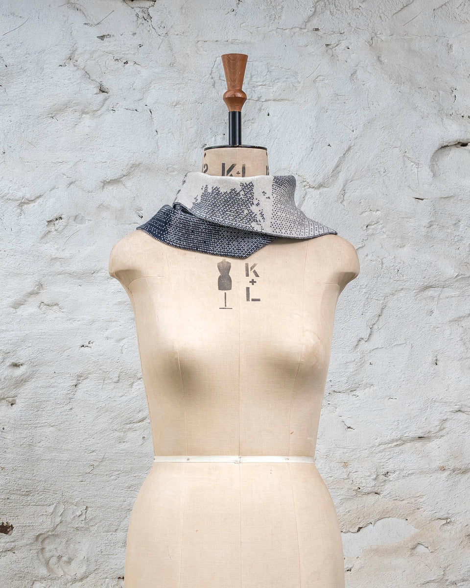Knitted Rani asymmetric cowl. Small abstract patterns make up a larger design with photographic imagery. Shown in inky blue and antique white