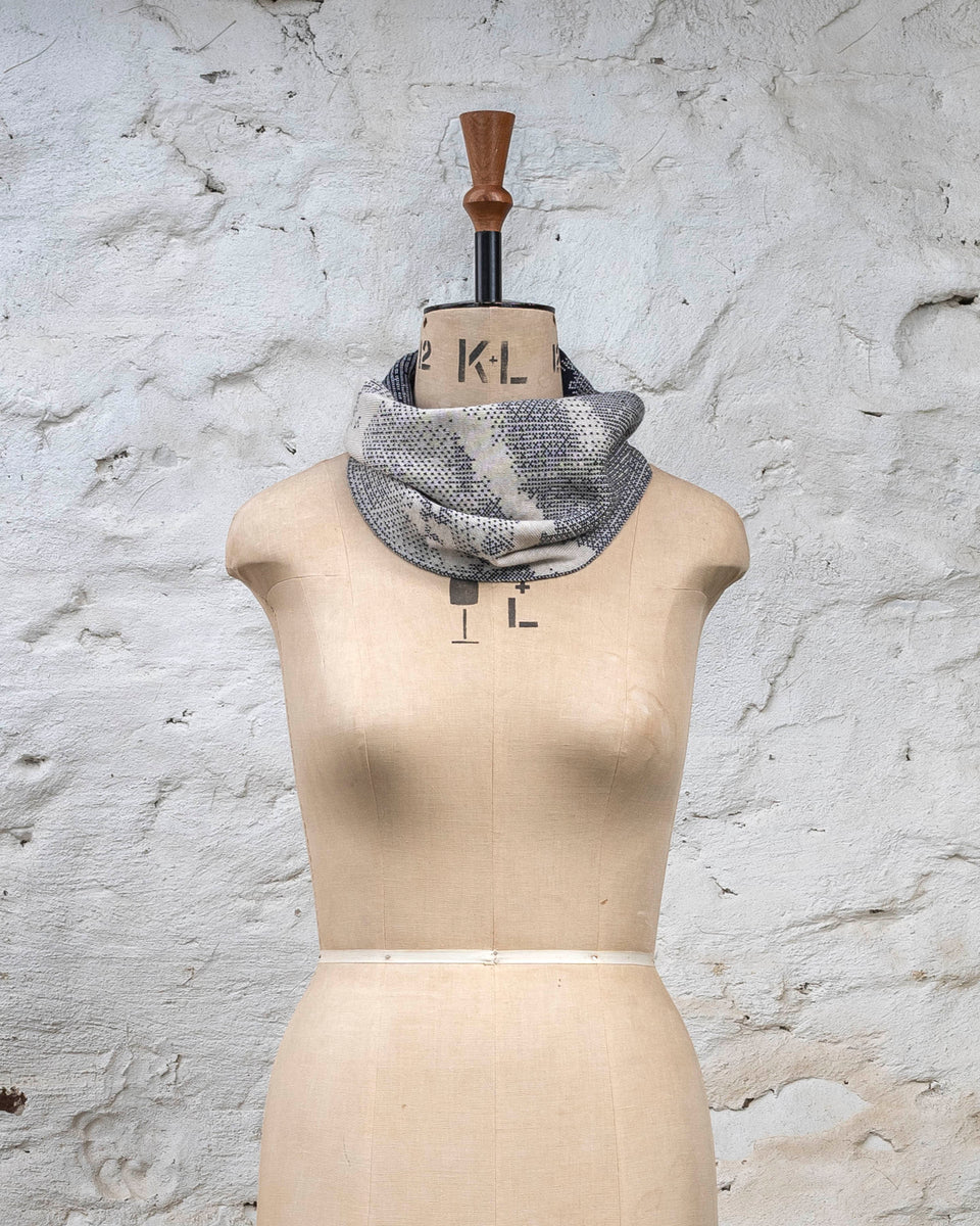 Knitted Rani asymmetric cowl. Small abstract patterns make up a larger design with photographic imagery. Shown in inky blue and antique white