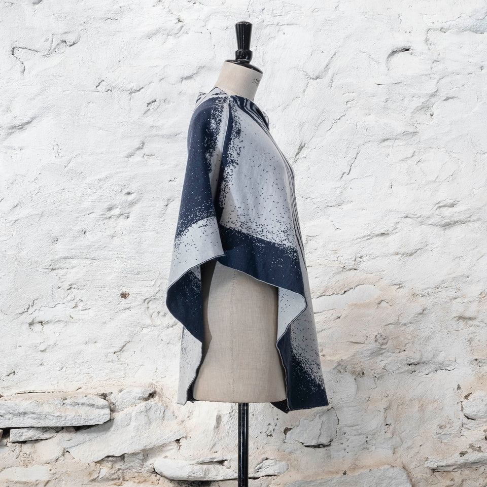 On a vintage dressmakers mannequin shown against a rustic whitewashed wall, a finely knitted poncho in a dark blue and off white. The pattern has an abstract, wavy linear design across one section of the cape. Shown from the left side, with panels of specked blue and white shown.
