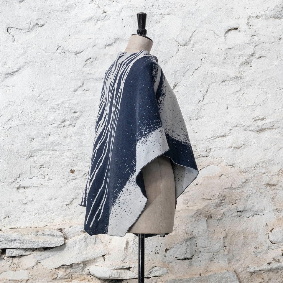 On a vintage dressmakers mannequin shown against a rustic whitewashed wall, a finely knitted poncho in a dark blue and off white. The pattern has an abstract, wavy linear design across one section of the cape. Shown from the left side, turned towards the back to see the pattern in reverse - the knit is double faced jacquard.