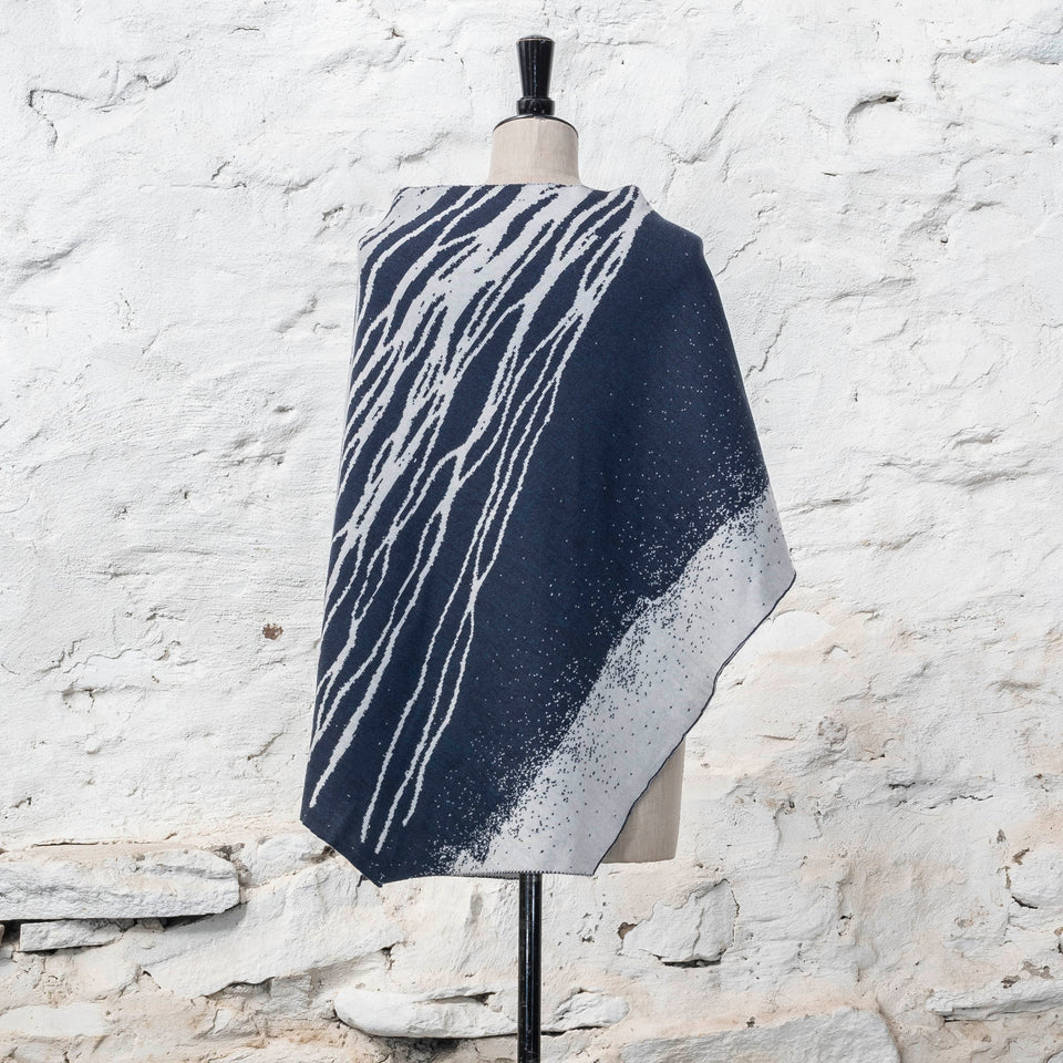 On a vintage dressmakers mannequin shown against a rustic whitewashed wall, a finely knitted poncho in a dark blue and off white. The pattern has an abstract, wavy linear design across one section of the cape. Shown from the back where the linear pattern is in reverse - white on blue - the knit is double faced jacquard.