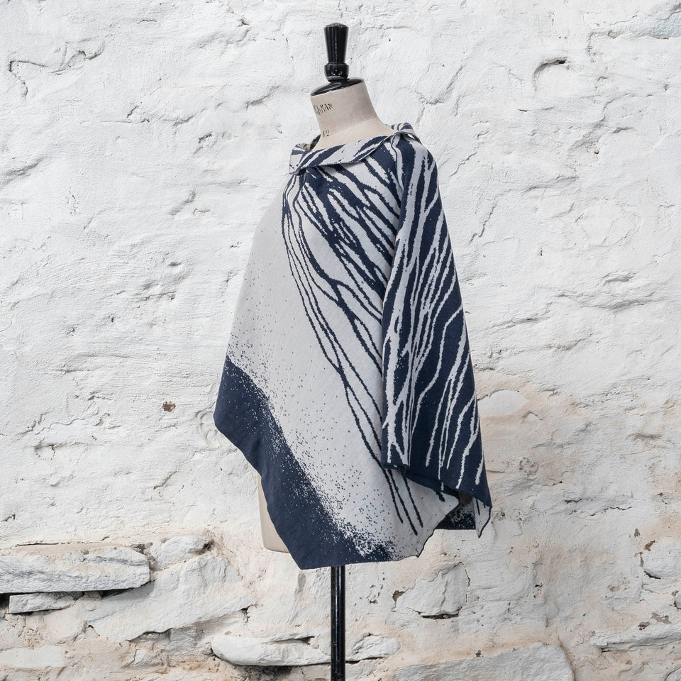On a vintage dressmakers mannequin shown against a rustic whitewashed wall, a finely knitted poncho in a dark blue and off white. The pattern has an abstract, wavy linear design across one section of the cape. Shown from the right, turned three-quarters. The linear pattern is shown with both secions reversed out at the shoulder. Blue on white and white on blue.
